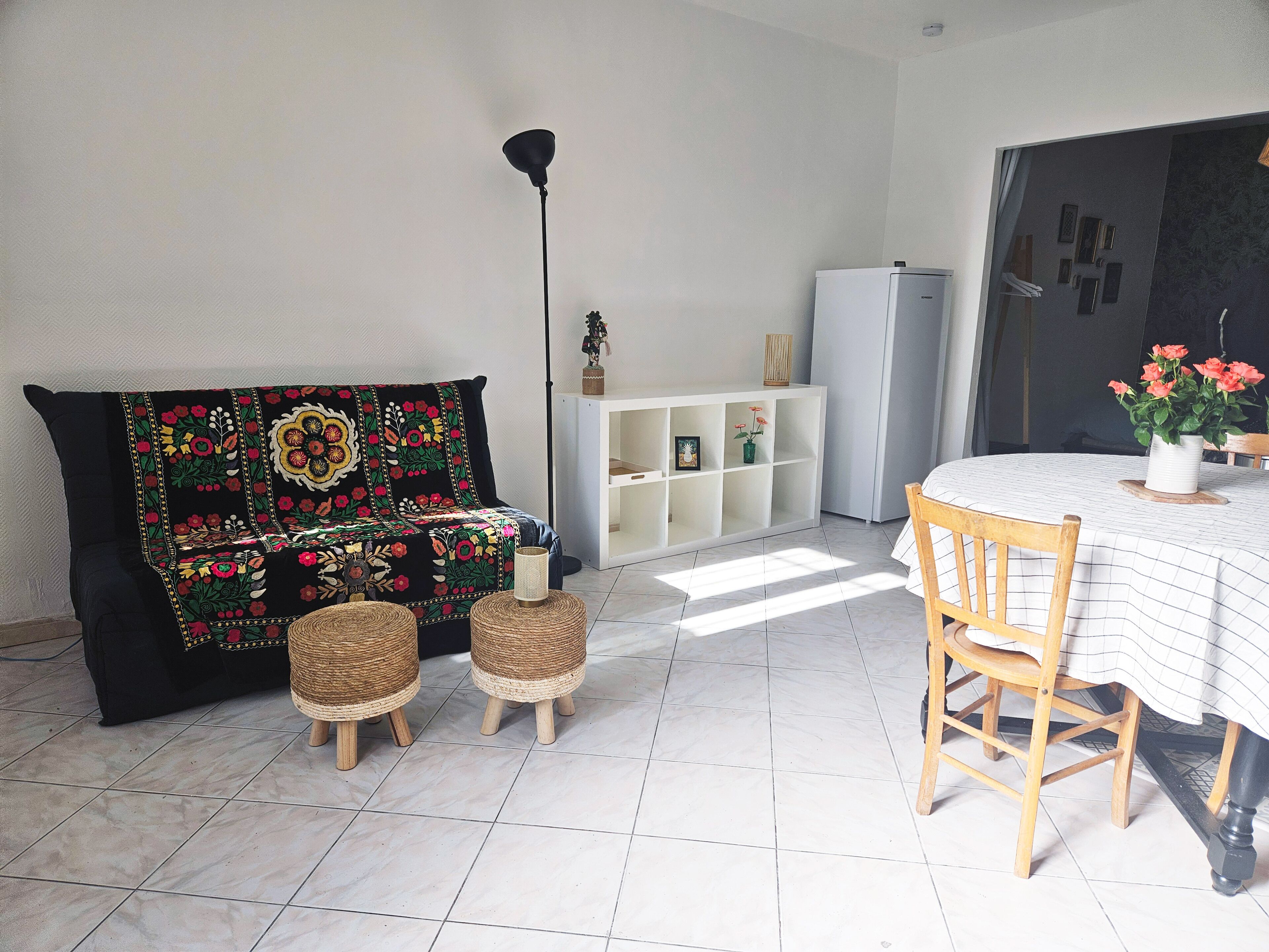 A living room with a sofa, two wicker stools, a dining table with a chair, a white cabinet, and a refrigerator.