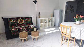 A living room with a sofa, two wicker stools, a dining table with a chair, a white cabinet, and a refrigerator.