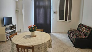 A dining area with a round table, chairs, a TV on a shelf, a flower vase, a sofa, and a window with curtains. - Apartment 40 m² near the Canal de la Robine (Narbonne)