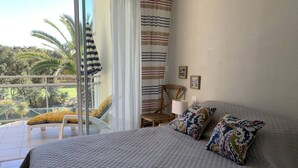 A bedroom with a large bed, a chair, a table, and a view of palm trees through a sliding glass door. - Résidence du Golf 223 - Moliets (MOLIETS ET MAA)