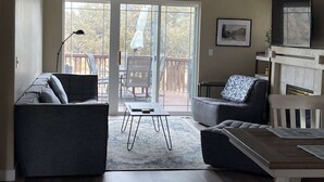 A modern living room with a sofa, armchair, and a fireplace. There is a sliding glass door leading to an outdoor area with a deck.