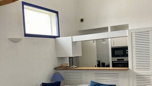 Un espacio habitable compacto con un sofá gris, una mesita de centro de vidrio, una pequeña zona de cocina con microondas y una ventana que proporciona luz natural.