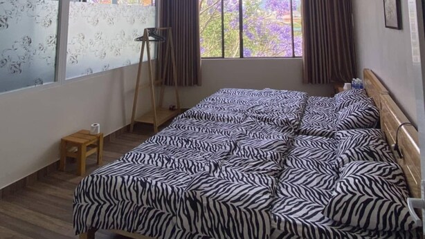 Quarto com uma cama grande, janela com cortinas, um banquinho de madeira e vista para árvores.