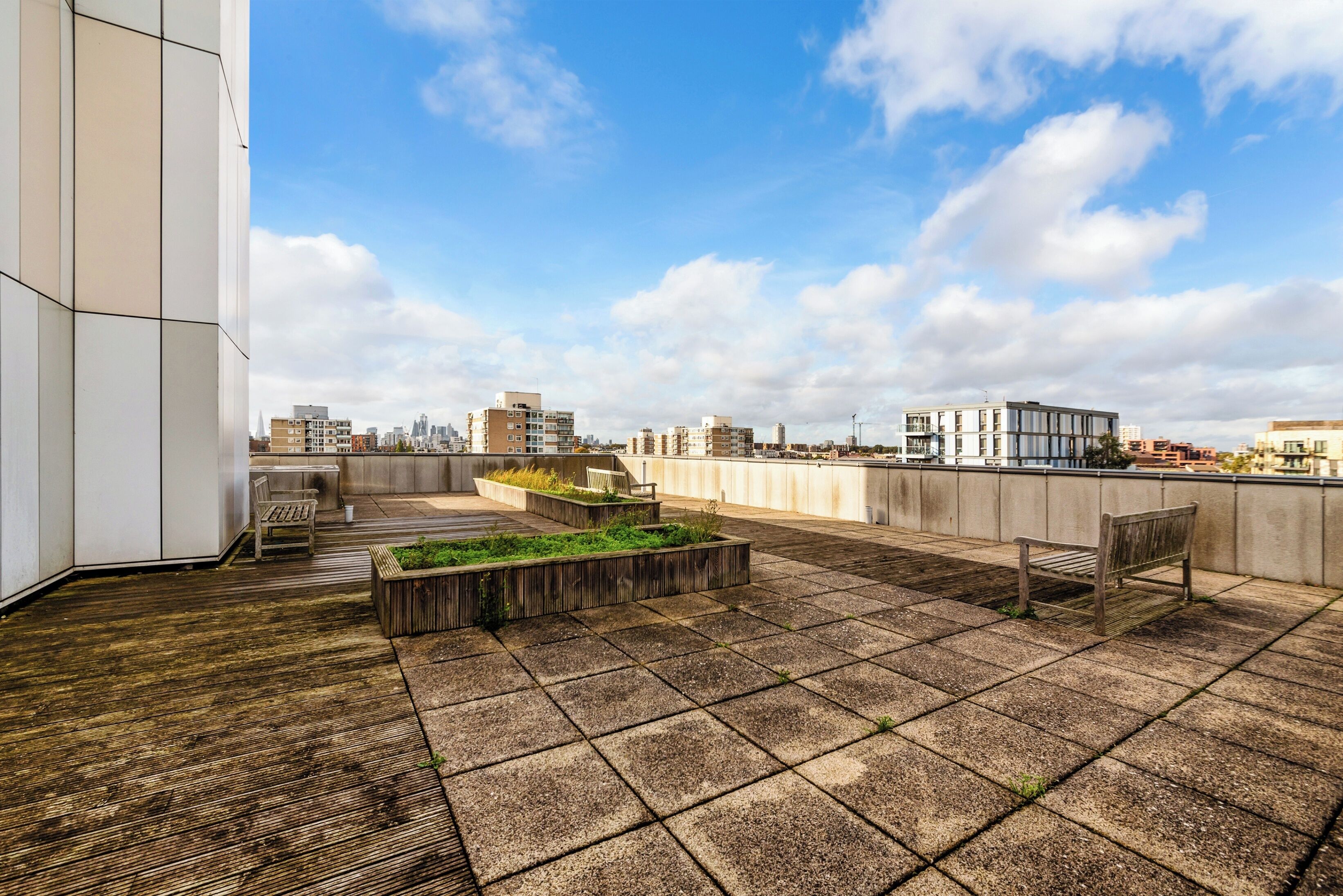 A rooftop terrace with a view of the city skyline, featuring a wooden bench, a planter with greenery, and a concrete wall.