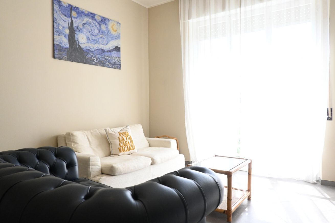 Een woonkamer met een zwarte bank, een witte bank, een schilderij aan de muur, en een raam met transparante gordijnen.