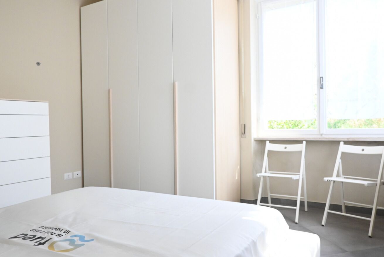 A bedroom with a bed, a wardrobe, two chairs, and a window.