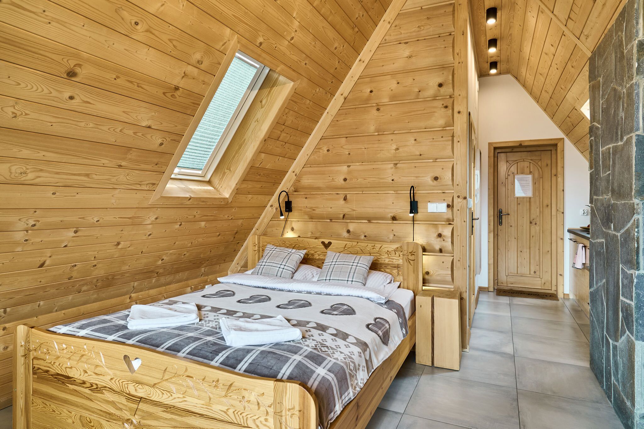 Ein Schlafzimmer einer Holzhütte mit schrägem Dach, einem Oberlicht, einem Bett mit gemustertem Überwurf, einem Nachttisch und einer Steinwand.