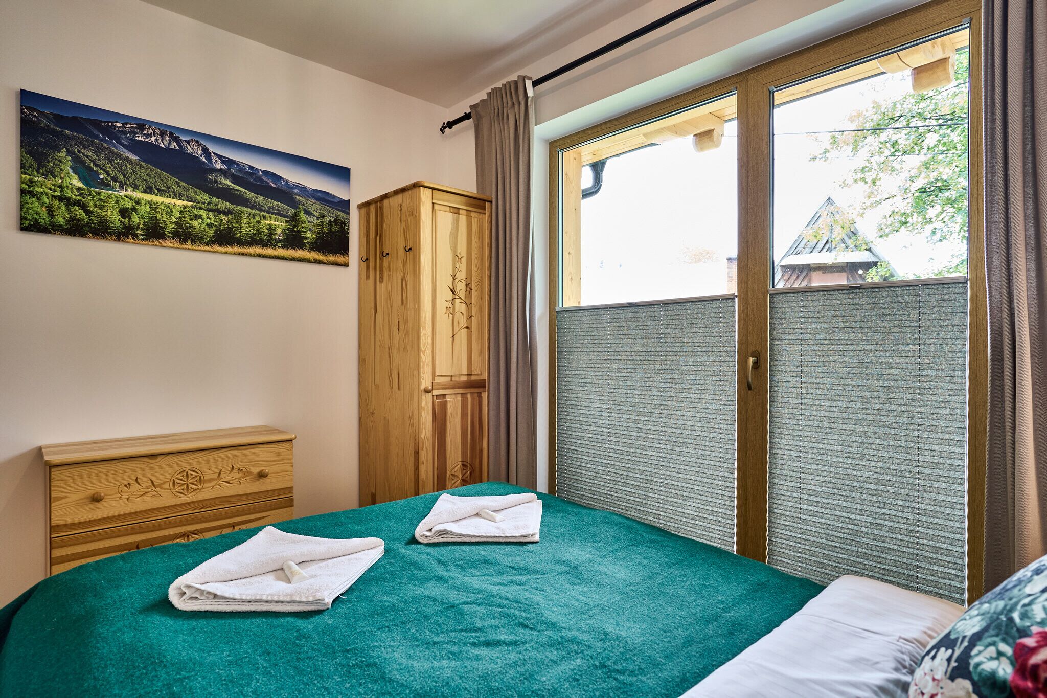 A bedroom with a bed, a wooden wardrobe, a window with a view of a mountain, and a painting on the wall.