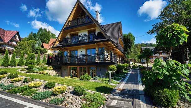 Ein zweistöckiges Haus mit Balkon, umgeben von gepflegten Gärten und blauem Himmel.