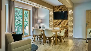 Ein modernes Wohnzimmer mit Holzbalkendecke, einer Essecke mit Tisch und Stühlen, einem an der Wand befestigten Flachbildfernseher und großen Fenstern mit Vorhängen.