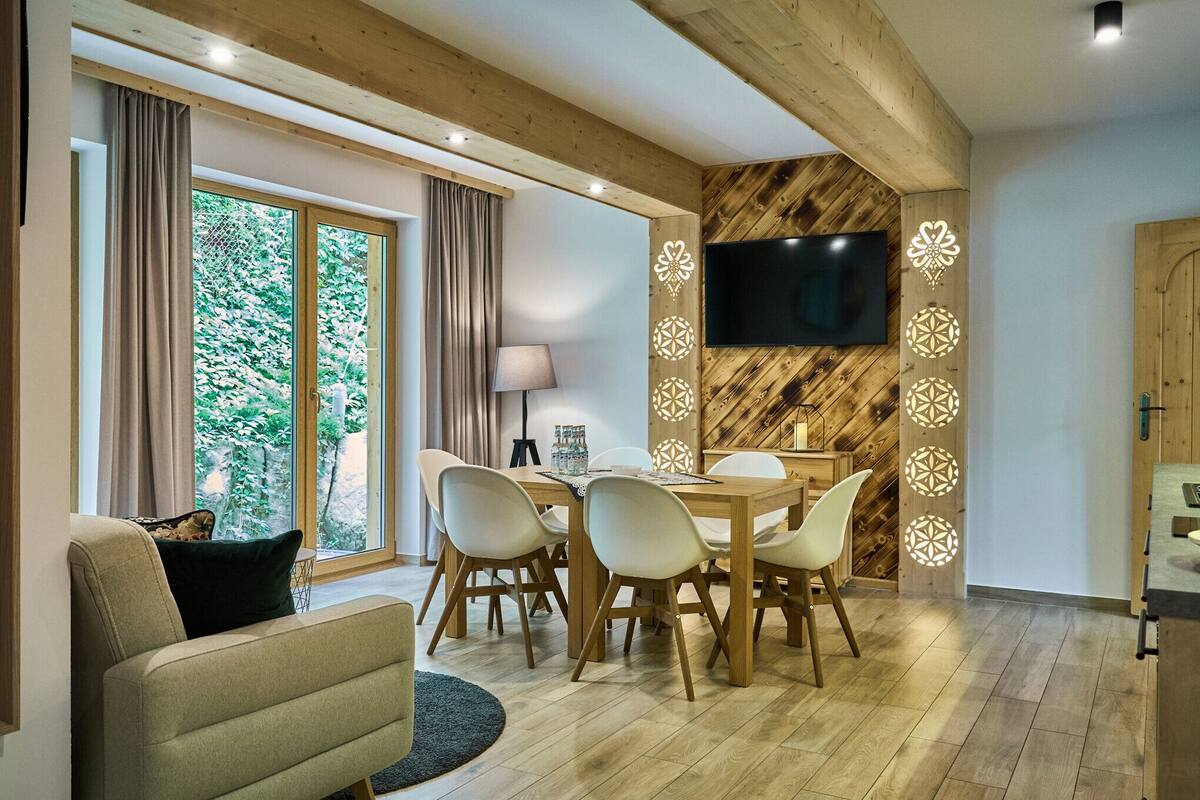 A modern living room with a wooden ceiling, a dining area with a table and chairs, a flat-screen TV mounted on the wall, and large windows with curtains.