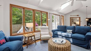 A sunlit living room with a ceiling fan, large windows, a white chair, a blue sofa, and a wicker ottoman.
