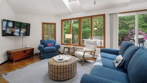 A spacious living room with a TV, sofas, a coffee table, and large windows offering a view of greenery.