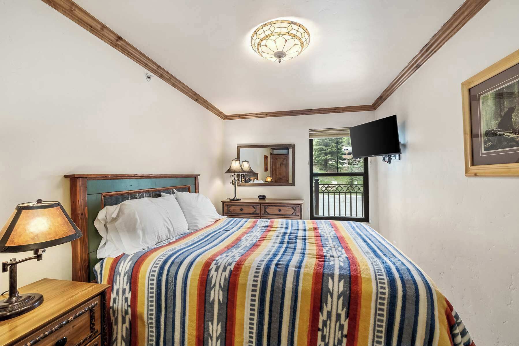 A bedroom with a bed, a nightstand, a lamp, a television, a mirror, and a window with a view.