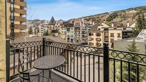 A balcony with a table and chairs, overlooking a town with various buildings and a hilly landscape.