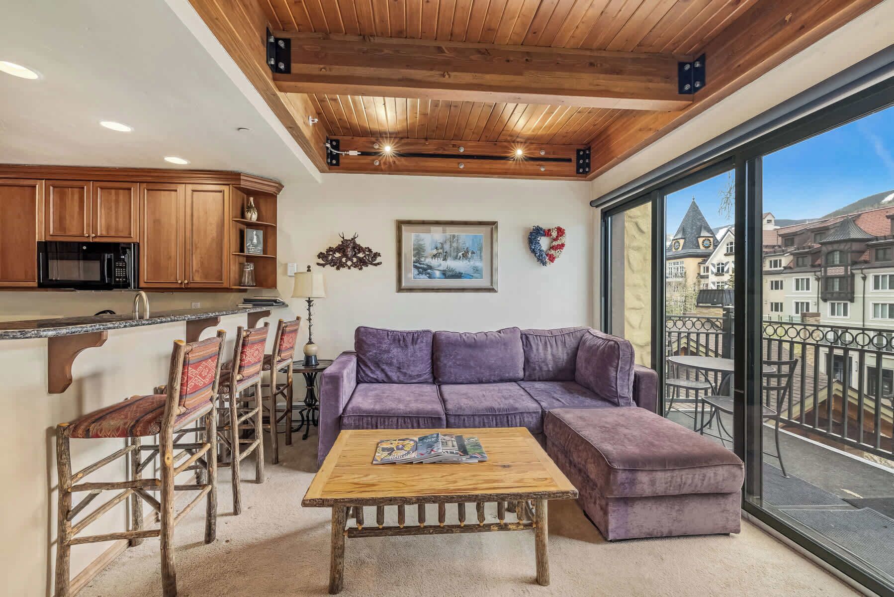 A modern living room with a purple sectional sofa, wooden coffee table, and dining chairs. A kitchen with wooden cabinets and a microwave is visible. A large window offers a view of buildings and a balcony.
