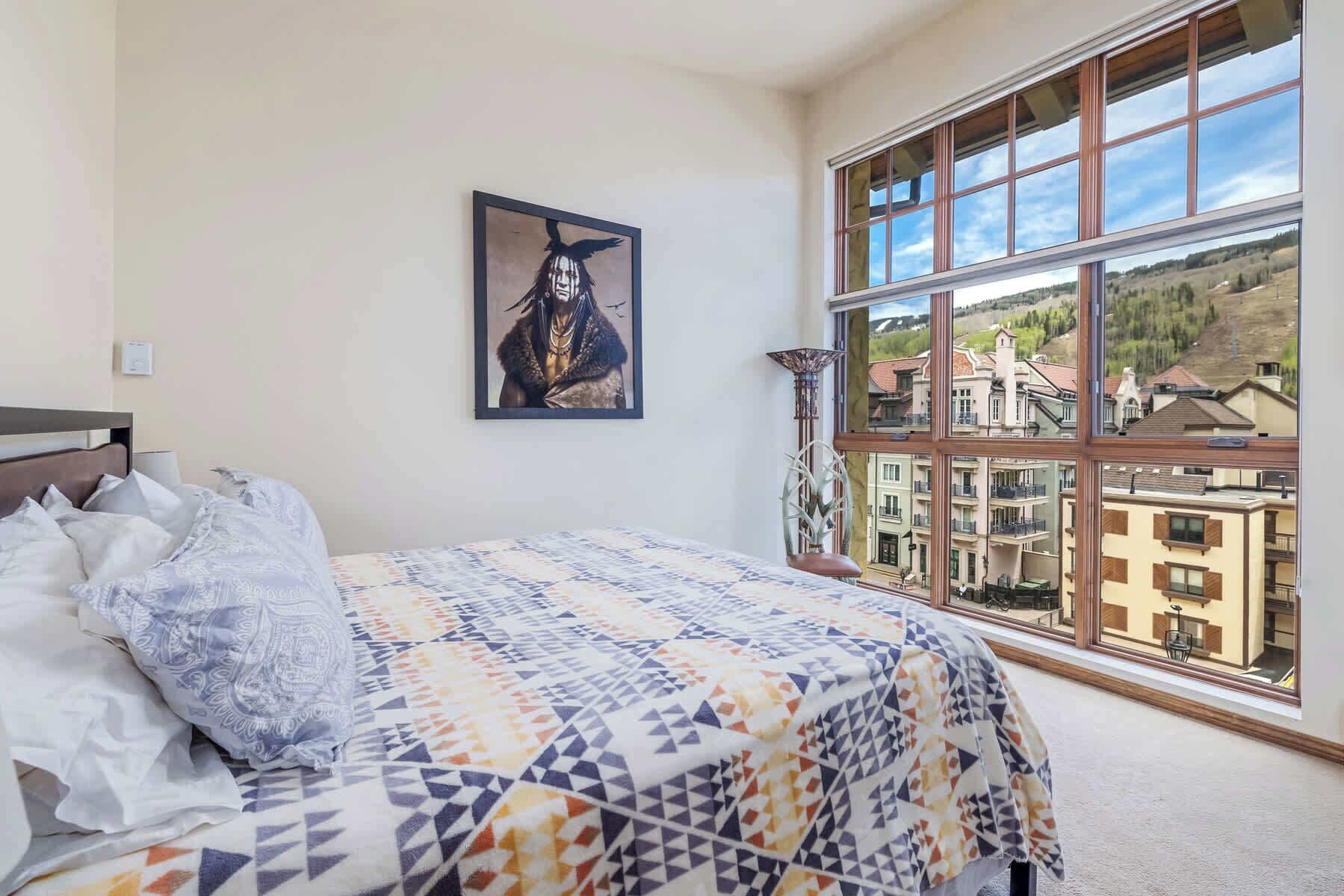 Een slaapkamer met een groot raam, een bed met beddengoed met een patroon en een ingelijst schilderij aan de muur.