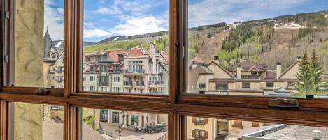 Vista a través de una ventana de un edificio de varios pisos, mostrando una combinación de edificios residenciales y comerciales, un cielo despejado y un paisaje montañoso al fondo.