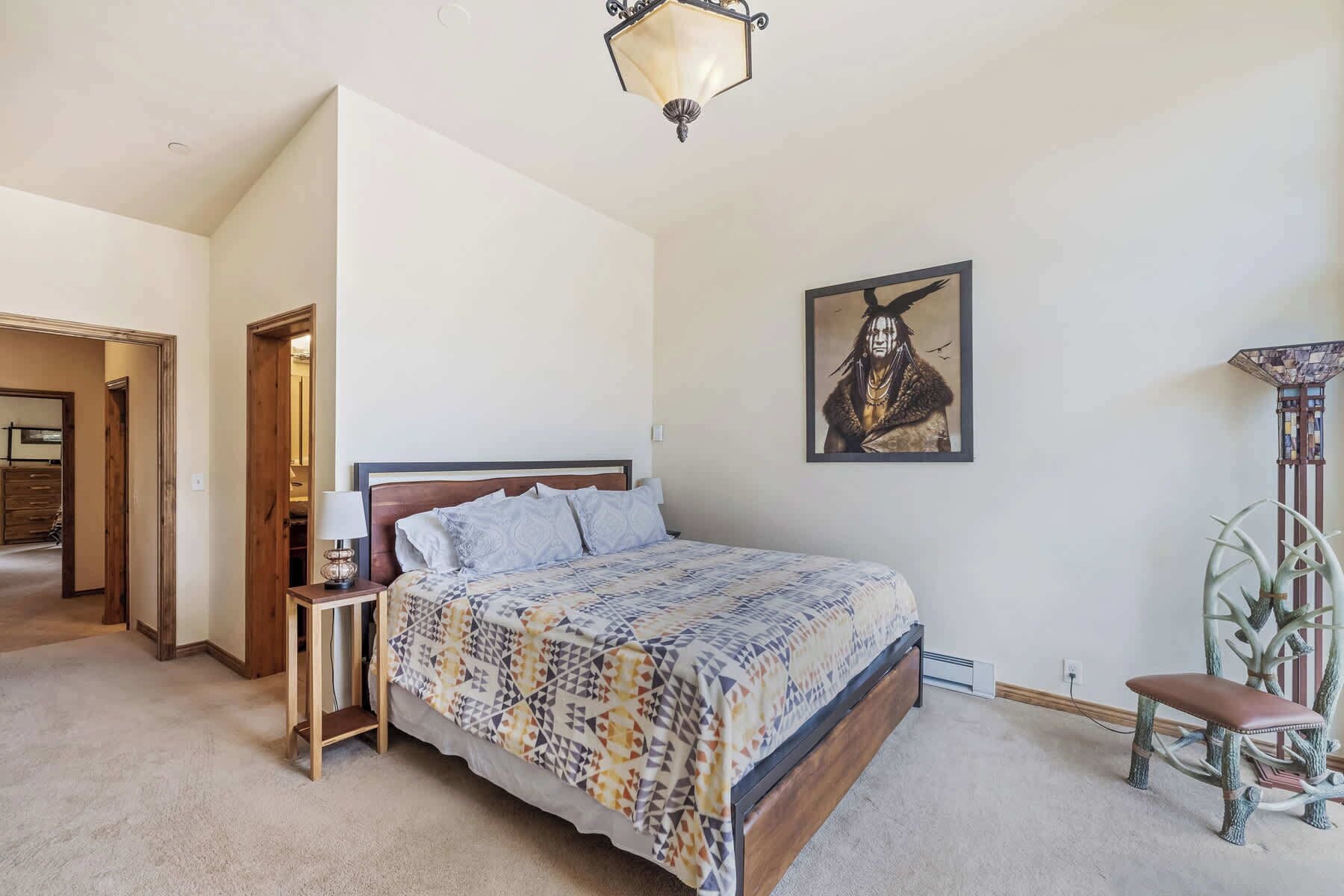 A bedroom with a bed, a nightstand, a lamp, a framed picture, a bench, and a decorative plant.