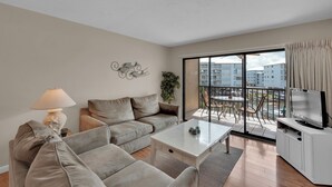 A modern living room with a sofa, a coffee table, a TV, and a balcony with a view of buildings. - 2B/2B ISLAND VACATION CONDO-RESORT POOL! WALK TO BEACH! SANTA MARIA 203 (Fort Myers Beach)