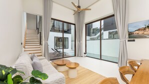 A modern living room with a sofa, coffee table, and dining table. There is a staircase leading to an upper level, and large windows with curtains. - Villa Qabalah (Si Sunthon)