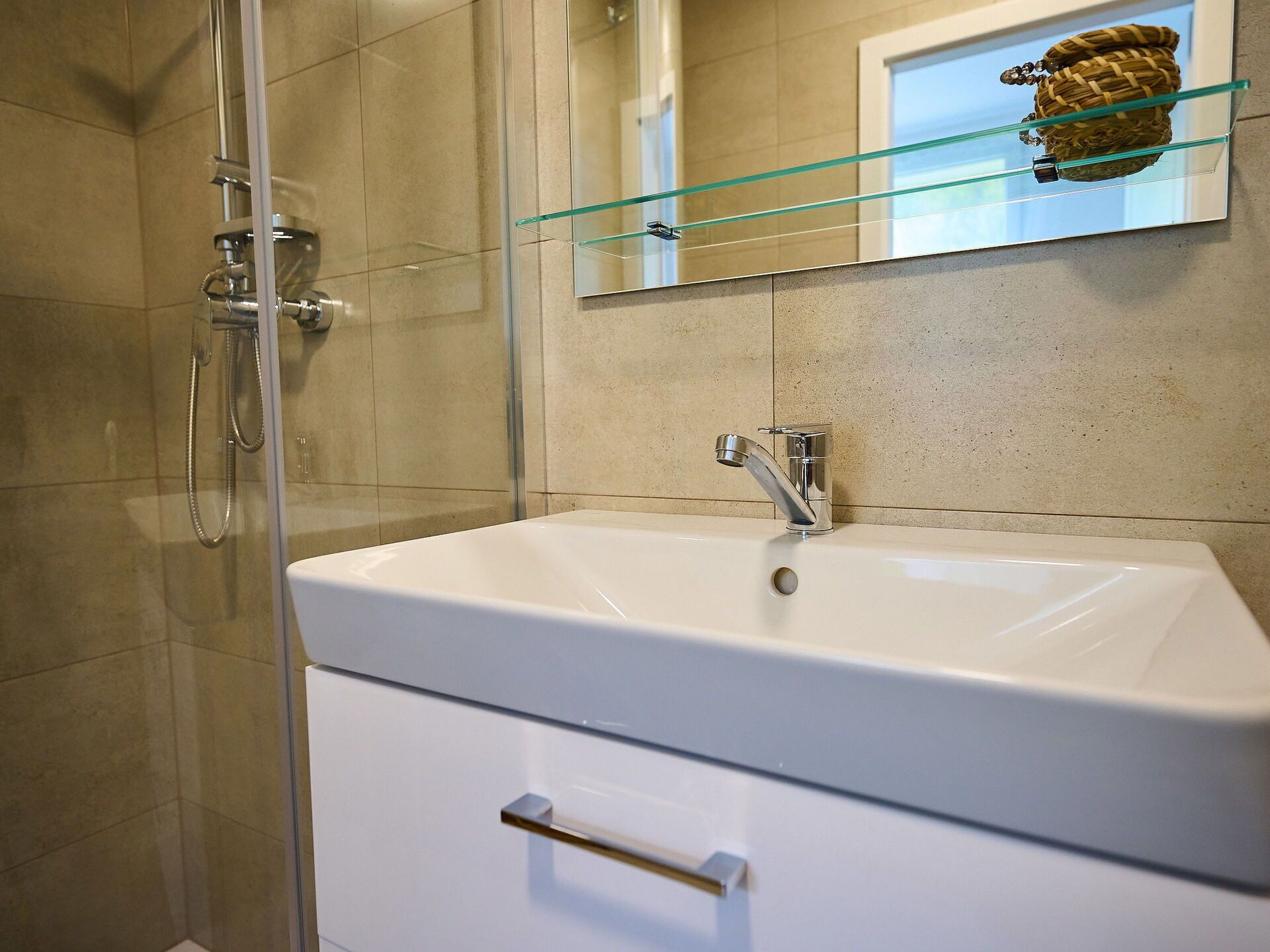 Een moderne badkamer met een glazen douchecabine, een witte wastafel met één kraan en een klein plankje boven de spiegel.