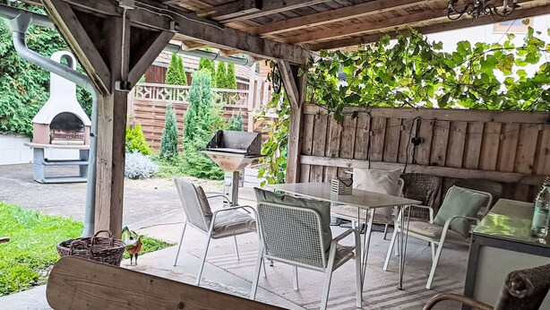 Ein Außenbereich mit einer Holzterrasse, einem Tisch, Stühlen, einem Grill und einer Pergola.