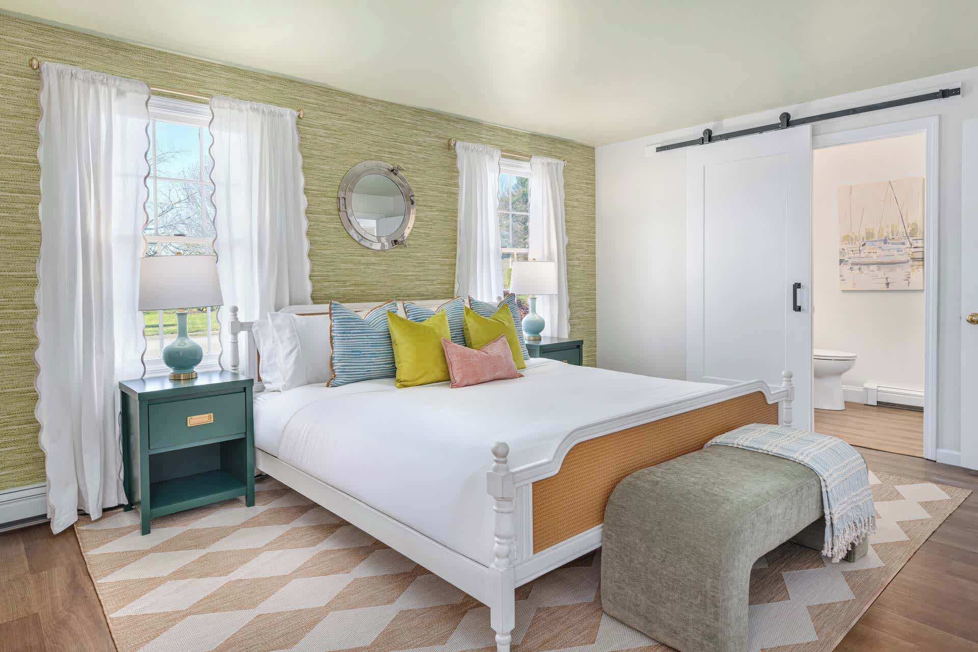 A bedroom with a bed, nightstands, a bench, a door, and a window with curtains.