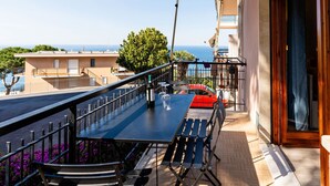 A balcony with a table and chairs, a red car parked in the background, and a view of the sea and buildings.