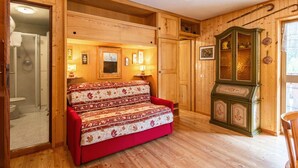 A cozy room with a wooden floor, a red sofa, a wooden cabinet with decorative items, and a shower area. - Nice apartment for 3 people with terrace (Canazei)