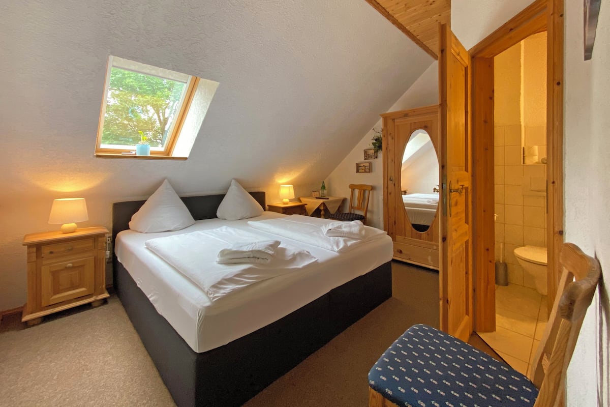 Une chambre avec un grand lit, un banc en bois, une table de chevet, une commode, un miroir et une fenêtre donnant sur des arbres.
