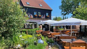 Un espace extérieur aménagé avec des tables et des bancs en bois, un petit étang et un bâtiment sur lequel est apposée une enseigne indiquant « QUERNWIRTSCHEFT/PENSION ».