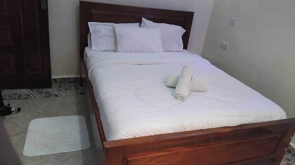A neatly made bed with white linens and a rolled towel on top, a wooden headboard, and a small white rug on the floor. - Lux Suites Grey Homes Nakuru (Nakuru)