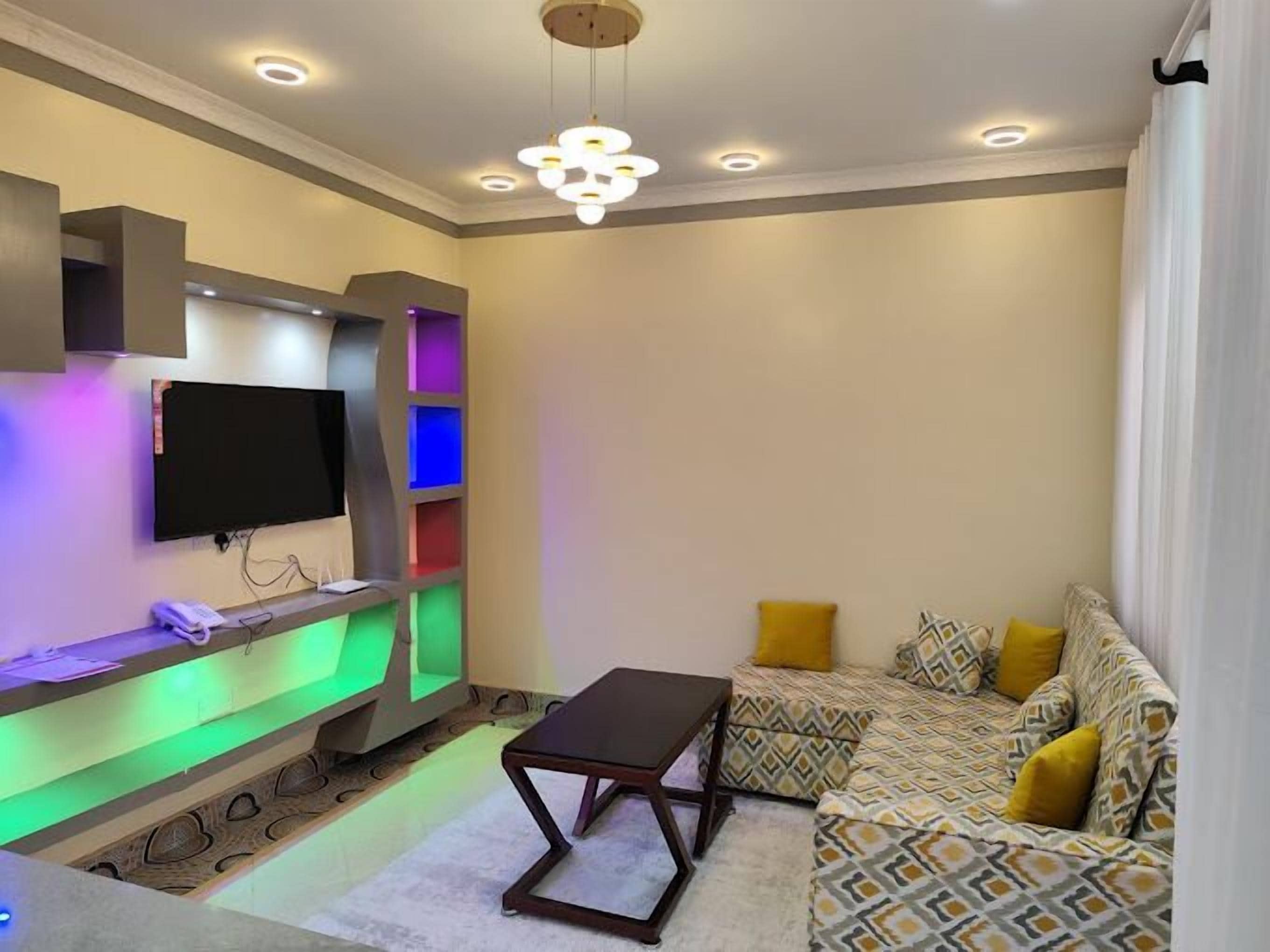 A modern living room with a flat-screen TV, a sofa with patterned cushions, a coffee table, and a wall-mounted entertainment unit with colorful lighting.