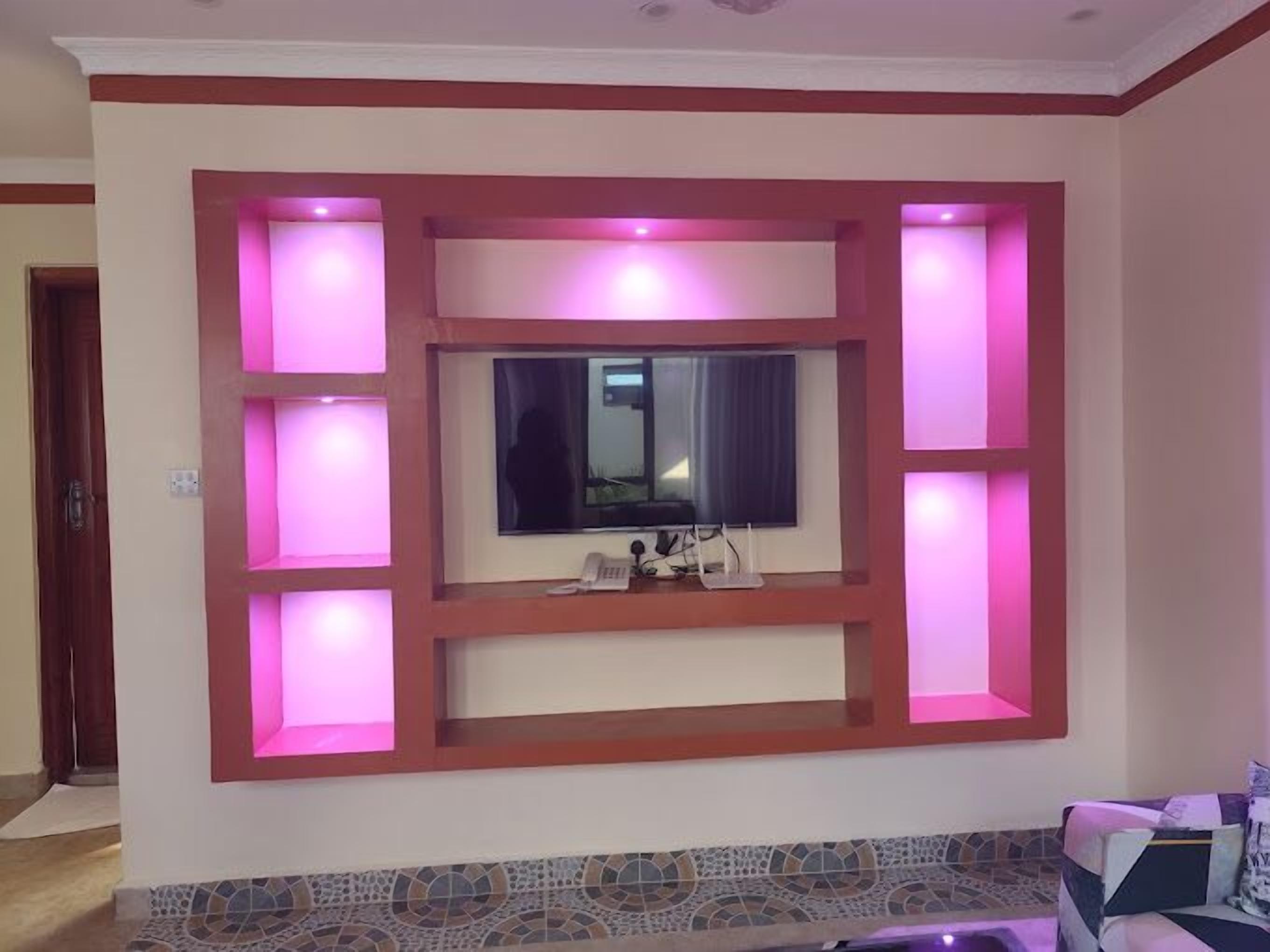 Ein Wohnzimmer mit an der Wand montiertem Fernseher, umgeben von eingebauten Regalen und rosarotem Licht.