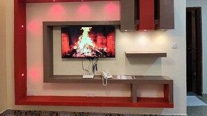 Een moderne woonkamer met een flatscreen televisie aan de muur, een inbouwbak eronder en een klein schap aan de rechterkant.