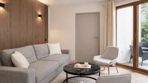 A modern living room with a grey sofa, a round coffee table, a white armchair, and a large sliding glass door leading to an outdoor area. - 1607 Brancion (Paris)