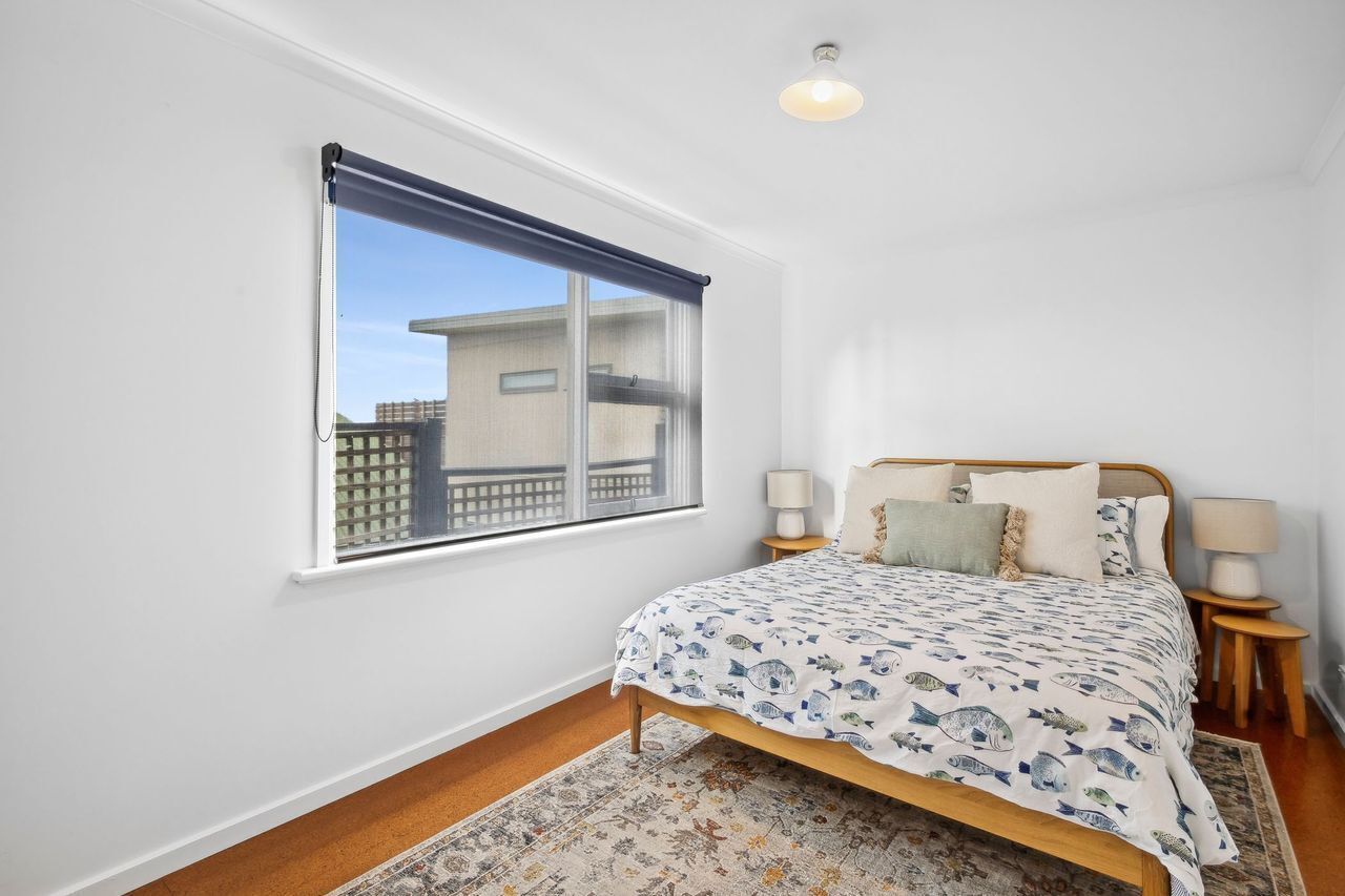 A bedroom with a bed, two bedside tables, a window with a blind, and a view of buildings outside.