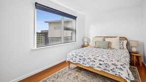 A bedroom with a bed, two bedside tables, a window with a blind, and a view of buildings outside.