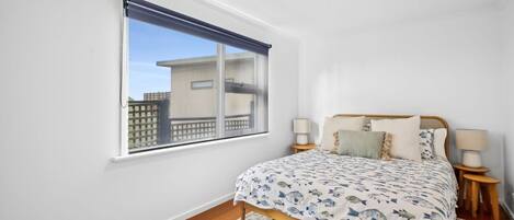 A bedroom with a bed, two bedside tables, a window with a blind, and a view of buildings outside.