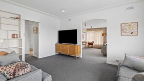 A living room with a flat-screen TV, grey sofas, a white bookshelf, and a dining area with a table and chairs.