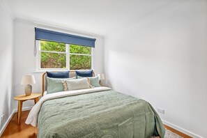 A bedroom with a bed, a window with a view of greenery, a bedside table with a lamp, and a power outlet on the wall.