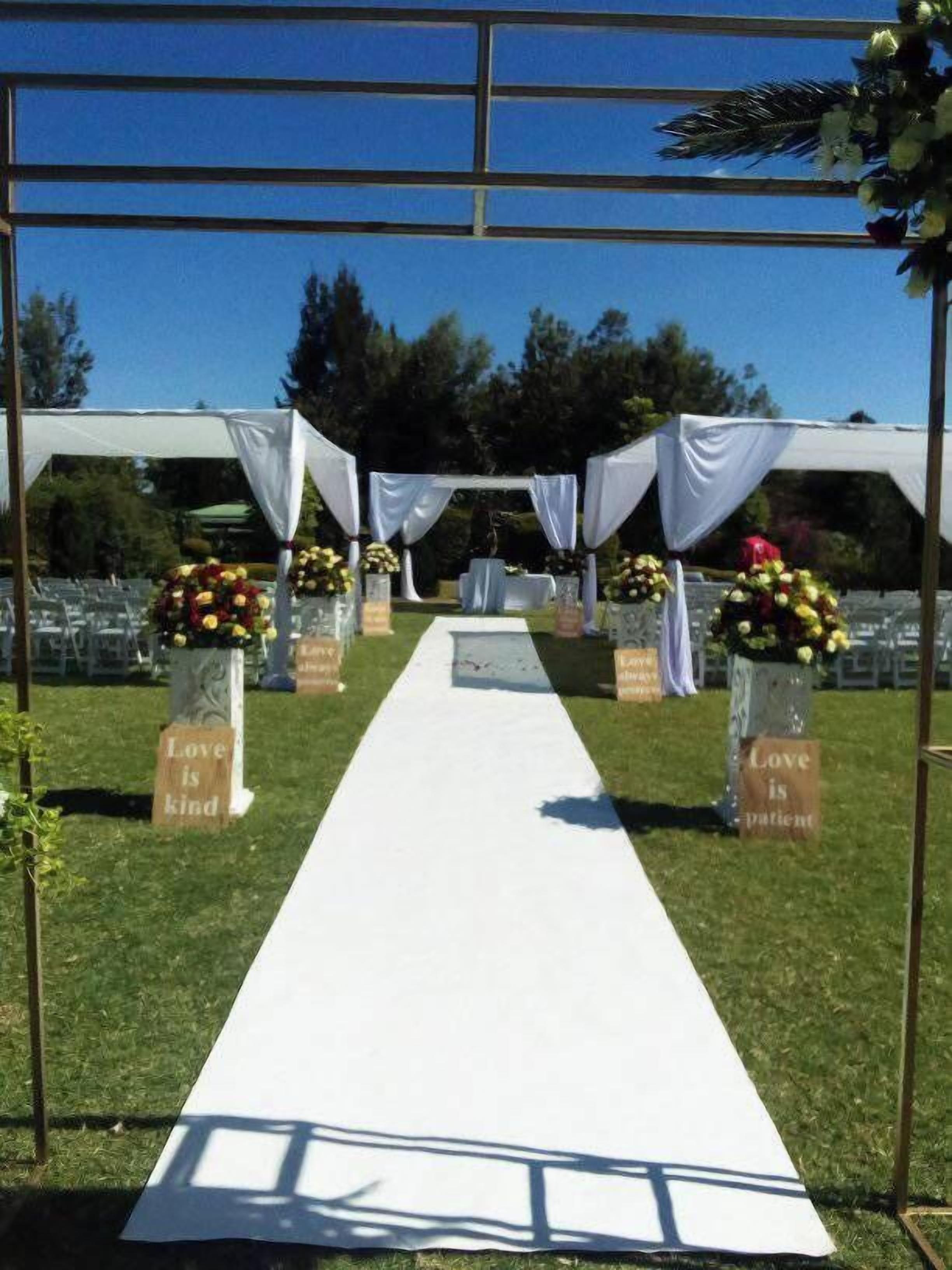 A white aisle leading to a canopy with white drapes, flanked by flower arrangements and signs stating "Love is kind" and "Love is patient."