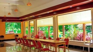 A conference room with a long table and red chairs, large windows with a view of greenery, and pendant lighting.