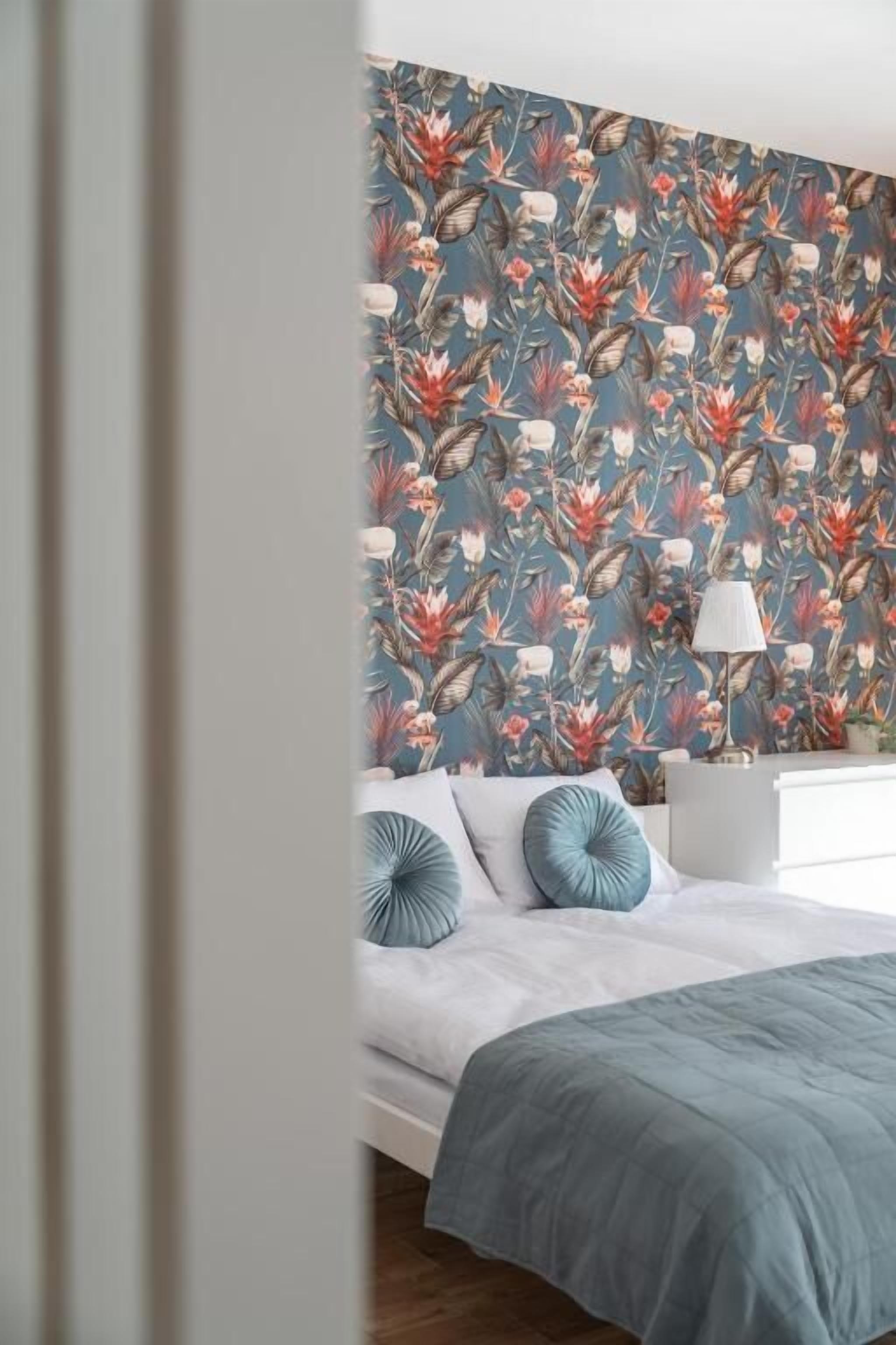 Ein Schlafzimmer mit floralem Wandpapier, einem Bett mit blauer Bettdecke und einem Nachttisch mit Lampe.
