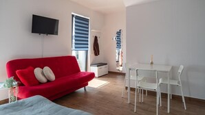 A modern room with a red sofa, white chairs, a TV mounted on the wall, and a window with blinds. - Przytulny apartament KOLEJOVA 12 (Nowy Targ)