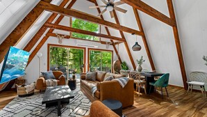 Um amplo loft com pé direito alto, vigas de madeira aparente, uma janela grande e um espaço de estar aconchegante com sofá e poltronas.