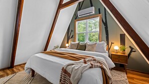 Um quarto com teto inclinado, uma cama com roupa de cama branca, uma mesa de cabeceira de madeira com abajur, e uma janela com vista para árvores.