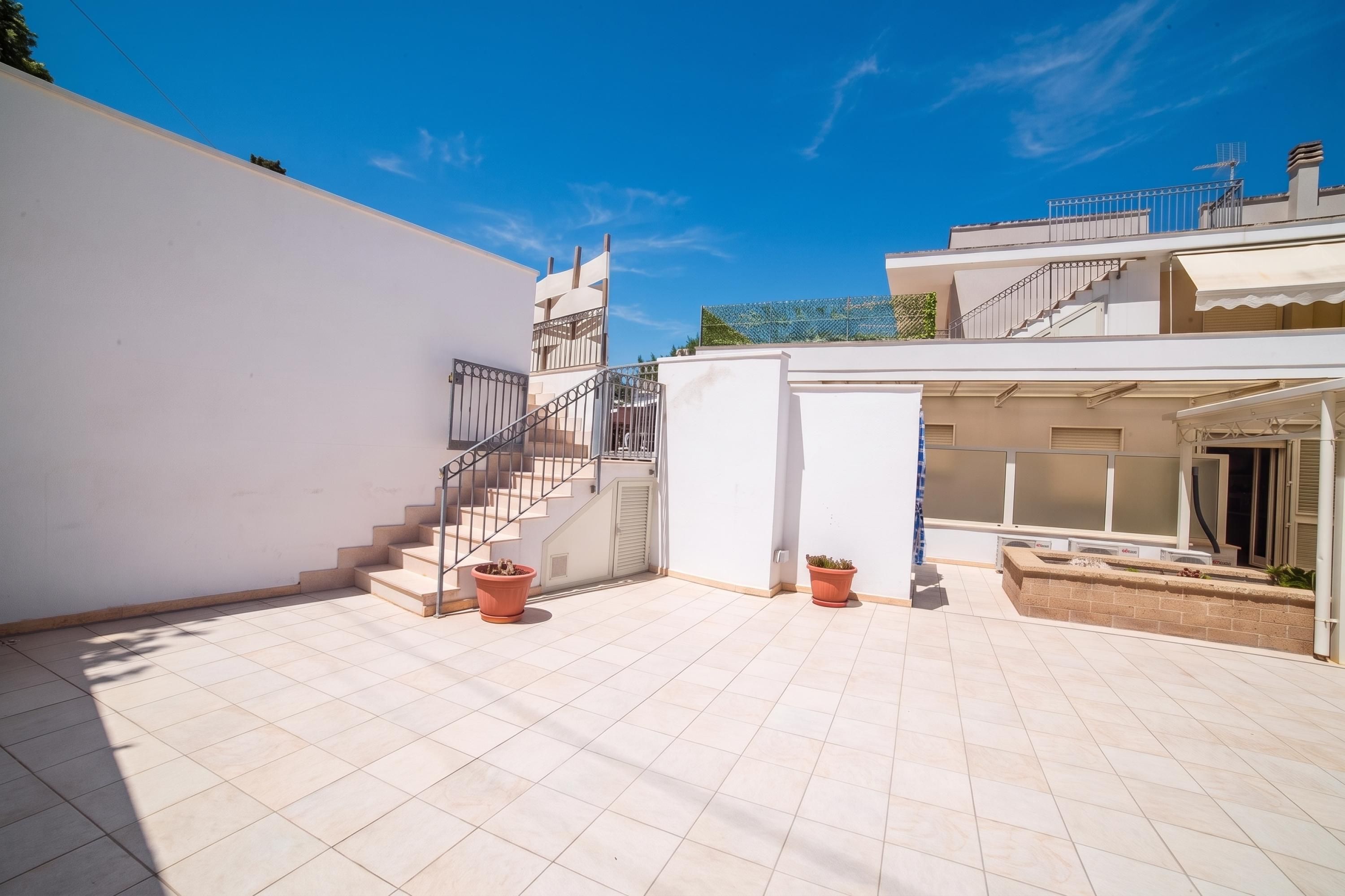 Un patio moderno al aire libre con una escalera que conduce a un balcón, plantas en macetas y un cielo azul despejado.