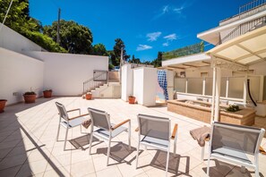 Une vaste terrasse extérieure avec un sol carrelé, plusieurs chaises, une petite table et un escalier menant à un niveau supérieur.